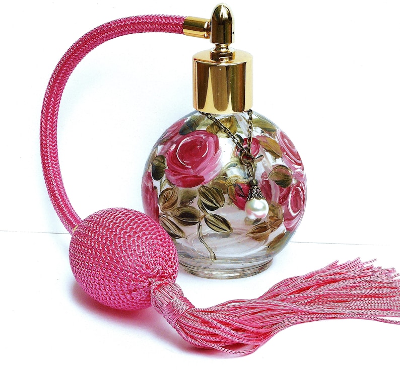 May include: A pink glass perfume bottle with a gold cap and a pink tassel. The bottle is decorated with hand-painted pink roses and green leaves. The bottle has a gold chain with a pearl charm.