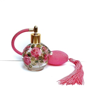 May include: A vintage glass perfume bottle with a pink tassel and a floral design. The bottle is clear glass with a gold top and a pink spray nozzle. The floral design features pink roses and green leaves.