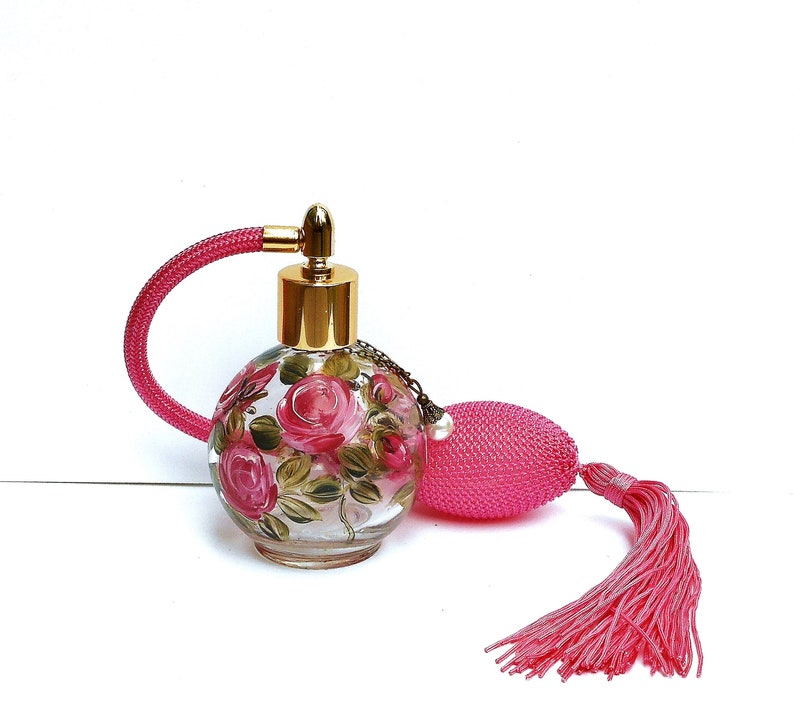 May include: A vintage glass perfume bottle with a pink tassel and a floral design. The bottle is clear glass with a gold top and a pink spray nozzle. The floral design features pink roses and green leaves.