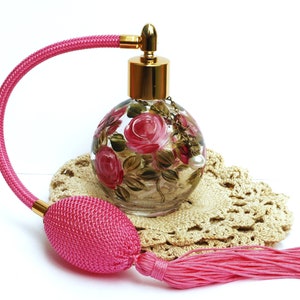 May include: A pink glass perfume bottle with a gold top and a pink tassel. The bottle is decorated with hand-painted pink roses and green leaves. The bottle is sitting on a crocheted doily.