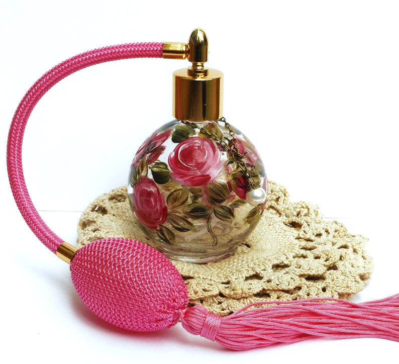 May include: A pink glass perfume bottle with a gold top and a pink tassel. The bottle is decorated with hand-painted pink roses and green leaves. The bottle is sitting on a crocheted doily.