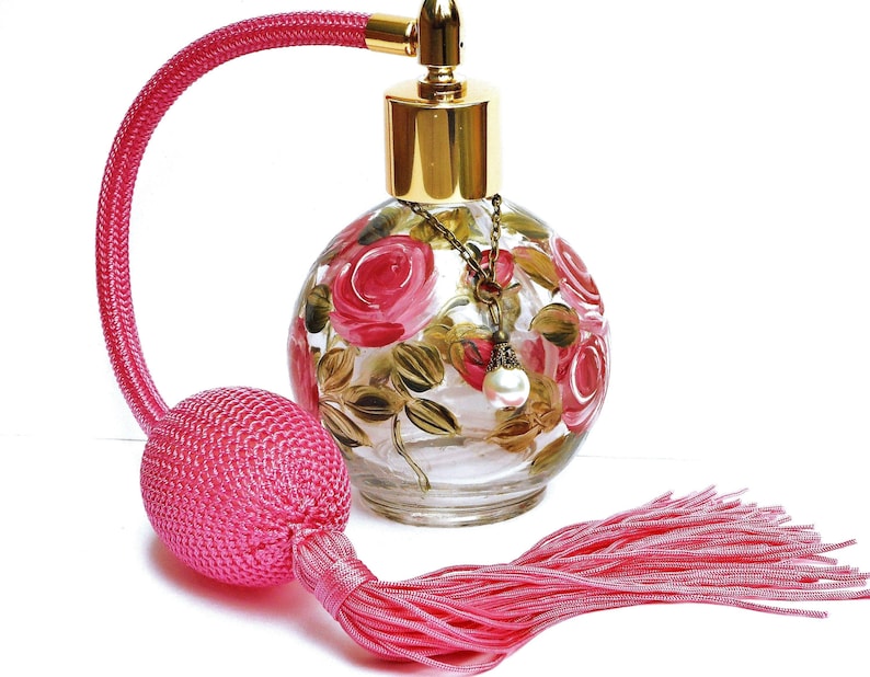 May include: A pink glass perfume bottle with a gold top and a pink tassel. The bottle is decorated with hand-painted pink roses and green leaves. The bottle has a gold chain with a pearl charm hanging from it.