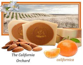 May include: Two bars of handmade soap with a brown centre and a white outer layer. The soap is labelled "californica". The image also shows almonds, a mandarin orange, and a slice of mandarin orange. The text "The California Orchard" is at the bottom of the image.