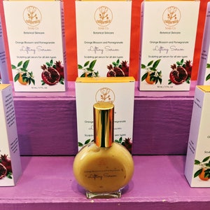 Puede incluir: Exhibici&oacute;n de productos para el cuidado de la piel. Una botella de vidrio transparente de Lifting Serum con una tapa dorada est&aacute; frente a varias cajas. Las cajas son blancas con detalles en naranja y rojo, y presentan el texto "Orange Blossom and Pomegranate Lifting Serum".