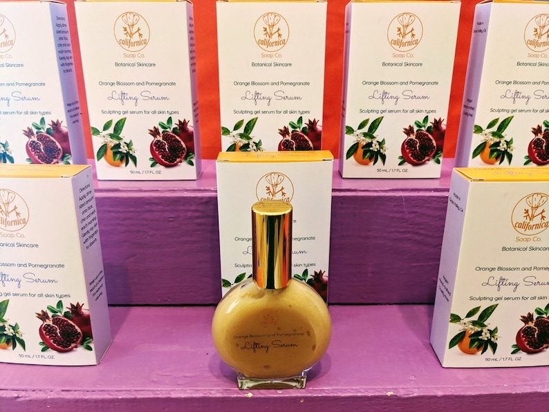 Puede incluir: Exhibici&oacute;n de productos para el cuidado de la piel. Una botella de vidrio transparente de Lifting Serum con una tapa dorada est&aacute; frente a varias cajas. Las cajas son blancas con detalles en naranja y rojo, y presentan el texto "Orange Blossom and Pomegranate Lifting Serum".