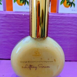 Puede incluir: Una botella redonda de vidrio transparente con una tapa dorada. La botella contiene un s&eacute;rum de color naranja claro con las palabras "Orange Blossom and Pomegranate Lifting Serum" impresas en la parte delantera. El fondo incluye cajas con ilustraciones de naranja y granada.