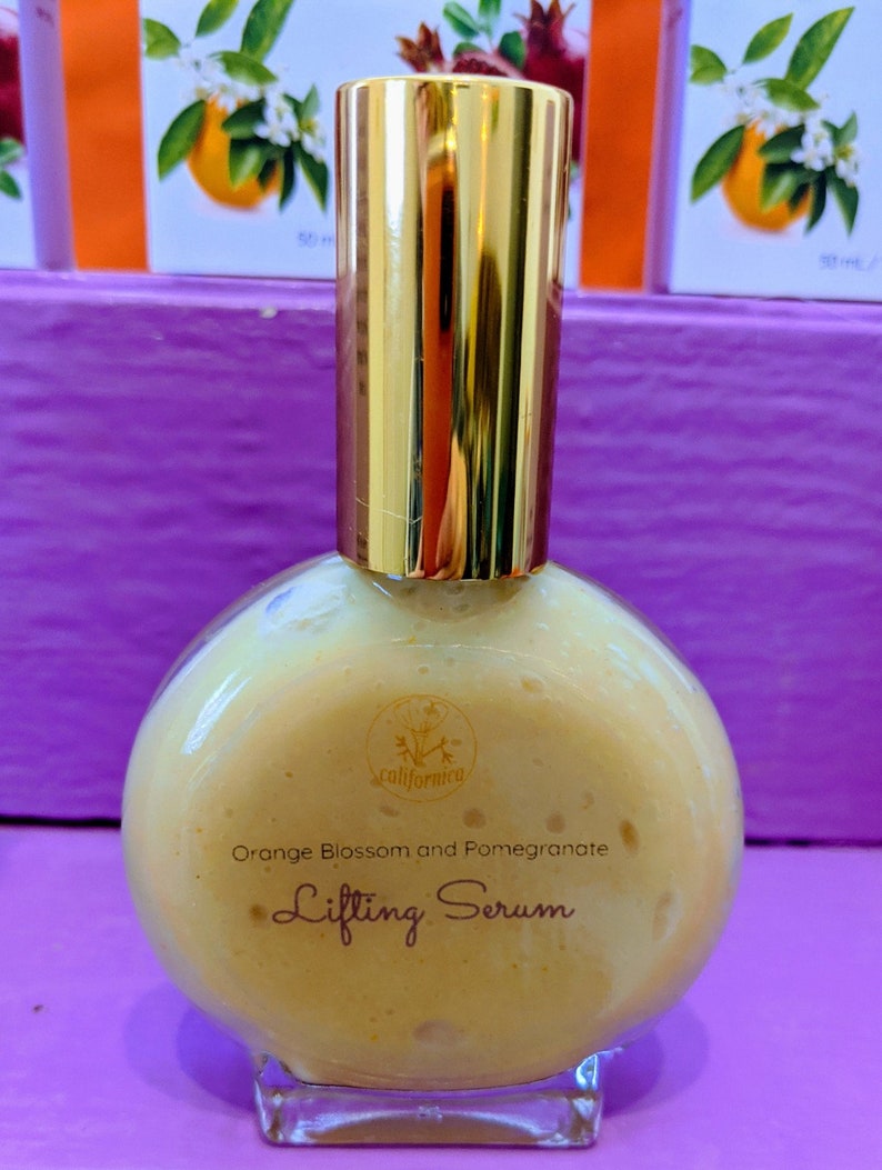 Puede incluir: Una botella redonda de vidrio transparente con una tapa dorada. La botella contiene un s&eacute;rum de color naranja claro con las palabras "Orange Blossom and Pomegranate Lifting Serum" impresas en la parte delantera. El fondo incluye cajas con ilustraciones de naranja y granada.