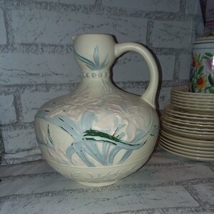 Può includere: Un'anfora in ceramica bianca con un motivo floreale in tonalità rosa, blu e verde. L'anfora ha una base larga e un collo stretto con un manico.