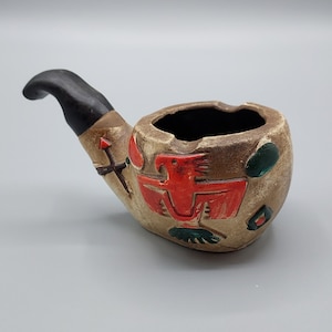 Könnte beinhalten: Ein Aschenbecher aus Keramik in Form einer Pfeife mit schwarzem Stiel. Die Schale der Pfeife ist mit einem roten, grünen und braunen Design verziert.
