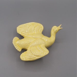 May include: A yellow ceramic bird figurine with its wings spread out in flight. The bird has a long neck and a small head with a beak.