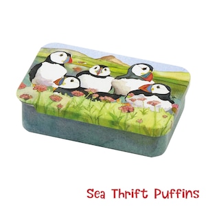 Könnte beinhalten: Eine rechteckige Blechdose mit einer Aquarellillustration von Papageientauchern auf einer Wiese mit rosa Blumen. Die Dose hat einen blauen Boden und einen Deckel mit dem Bild. Der Text unten lautet "Sea Thrift Puffins."