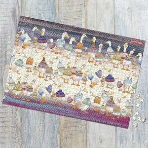 May include: A completed jigsaw puzzle featuring a whimsical illustration of numerous ducks wearing colorful sweaters and hats. The puzzle has a knitted border in shades of purple, blue, and gray. The artist's name, Emma Ball, is visible in the upper right corner.