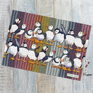 May include: A completed jigsaw puzzle featuring a colorful illustration of puffins. The puzzle pieces are assembled to show multiple puffins with white, black, and orange coloring, set against a striped background. The artist's name, "Emma Ball", is visible.