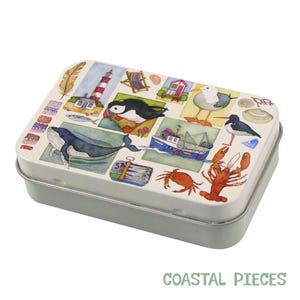 Puede incluir: Una lata rectangular con base verde claro y tapa color crema. La tapa presenta un collage de ilustraciones costeras, incluyendo una ballena, frailecillo, faro y langosta. El texto "COASTAL PIECES" está impreso en la parte inferior.