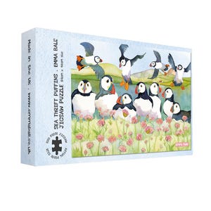 May include: A 1000-piece jigsaw puzzle featuring a watercolor illustration of puffins in a coastal scene. The puzzle box displays the text "SEA THRIFT PUFFINS" and measures 64cm x 48cm. The puzzle is made in the UK.