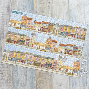 May include: A completed jigsaw puzzle featuring a colorful illustration of a street scene with buildings, shops, and yellow cars. The puzzle has a light blue sky background and the name "Emma Ballo" is visible in the upper left corner. Some puzzle pieces are separate.