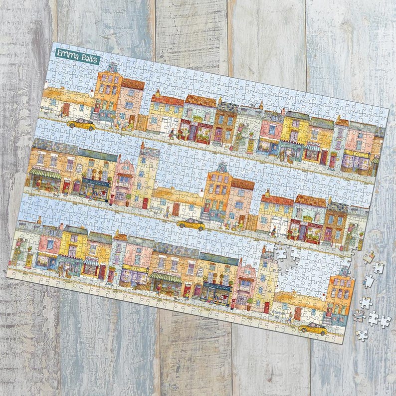 May include: A completed jigsaw puzzle featuring a colorful illustration of a street scene with buildings, shops, and yellow cars. The puzzle has a light blue sky background and the name "Emma Ballo" is visible in the upper left corner. Some puzzle pieces are separate.