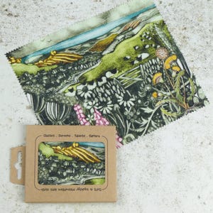 May include: A small, square, brown cardboard box with a clear plastic window showing a colorful printed microfiber cleaning cloth. The cloth features a landscape scene with green hills, a blue sky, and yellow flowers. The box has the text "Glasses, Screens, Tablets, Camera" and "Soft & Absorbent Microfibre Lens Cloth" printed on it.