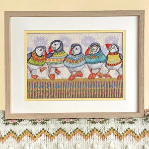 May include: A framed cross-stitch artwork featuring five colorful puffins in sweaters. The puffins are standing in a row, with a background of a fence and a light purple sky. The frame is a light wood color.