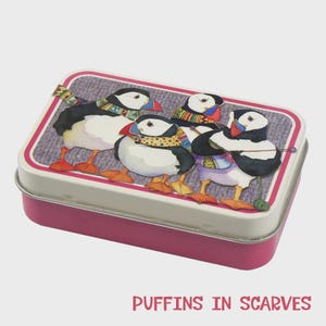 Puede incluir: Una lata rectangular con base rosa y tapa blanca con un diseño de cuatro frailecillos con bufandas de colores. Los frailecillos tienen plumas blancas y negras, patas naranjas y picos rojos. El texto "PUFFINS IN SCARVES" está impreso en la parte inferior.