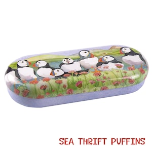 May include: A purple and white oval-shaped glasses case with a colourful illustration of puffins on a grassy cliffside. The text "SEA THRIFT PUFFINS" is printed on the case.