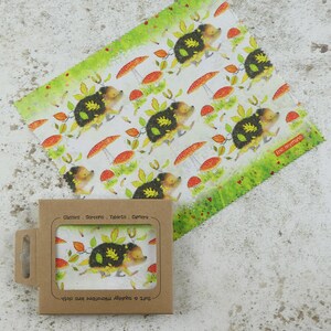 May include: A brown cardboard box with a clear plastic window showing a microfiber cleaning cloth with a hedgehog design. The cloth has a white background with red mushrooms, green leaves, and brown hedgehogs. The text on the box reads "Glasses - Screens - Tablets - Camera" and "Soft & Absorbent Microfibre Lens Cloth".