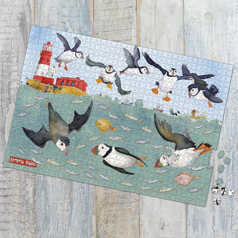 May include: A completed jigsaw puzzle featuring a whimsical illustration of puffins in flight over the sea. The puzzle includes a lighthouse, fish, and boats. The puzzle is by Emma Ball.