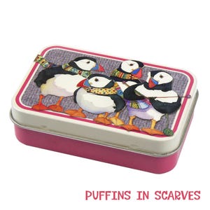 May include: A rectangular metal tin with a pink base and a white lid featuring an illustration of three puffins wearing colourful patterned scarves. The puffins have black and white feathers, orange feet, and multi-coloured beaks. The text reads PUFFINS IN SCARVES.