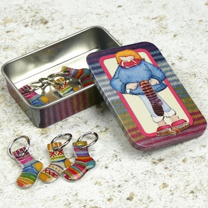 May include: A set of colourful sock stitch markers in a tin with a cartoon illustration of a person knitting a sock. The tin has a pink, blue and green plaid pattern.