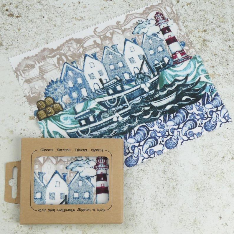 May include: A set of two microfiber cleaning cloths featuring a blue and white illustration of a lighthouse, houses, and a boat. The text "Glasses - Screens - Tablets - Camera" is printed on the packaging.