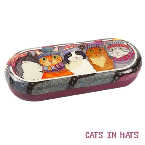 May include: A purple and gray oval-shaped glasses case with a design of five cats wearing hats. The cats are wearing colorful hats and scarves. The case has the words "CATS IN HATS" printed on the bottom.