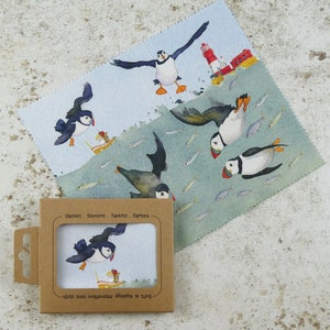 Puede incluir: Dos paños de limpieza de microfibra con un fondo azul y verde que presenta un patrón repetitivo de frailecillos y peces. Los paños están empaquetados en una caja de cartón marrón con el texto "Soft & Absorbent Microfibre Lens Cloth" y "Glasses - Screens - Tablets - Camera".