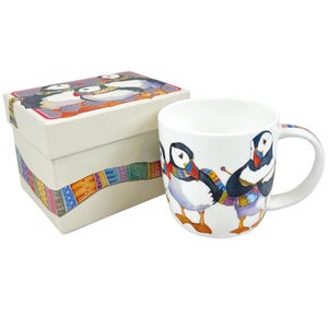 Op de afbeelding: Witte keramische mok met een kleurrijke illustratie van papegaaiduikers met sjaals. De mok wordt gepresenteerd met een bijpassende geschenkdoos. De papegaaiduikers hebben zwart-witte veren, oranje snavels en zijn aan het breien.