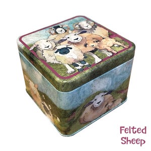 Könnte beinhalten: Eine quadratische Blechdose mit einer bunten Illustration von Schafen auf einer grünen Wiese. Die Dose hat einen rosa Rand und die Worte "Felted Sheep" sind am unteren Rand des Bildes aufgedruckt.