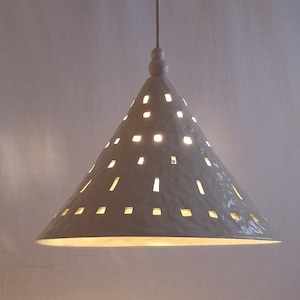 May include: White ceramic pendant light with a cone-shaped shade featuring a pattern of small, square cutouts. The light is turned on, illuminating the cutouts.