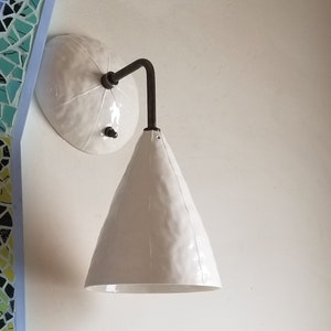 May include: White ceramic wall sconce with a textured finish. The sconce features a conical shade and a circular backplate, connected by a dark metal arm. The fixture is mounted on a white wall.