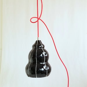 Black ceramic pendant light has an organic form and is detailed with thin white stripes. Hangs from a red 15' plugin cord