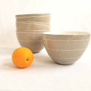 stack of porcelain serving bowls in taupe with thin white stripes. Shown with an orange for scale