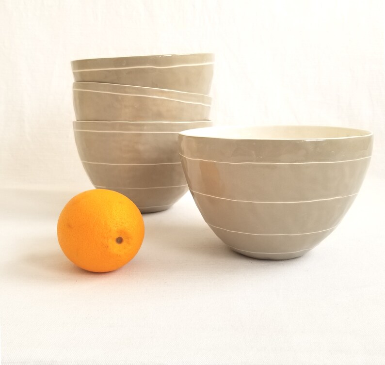 stack of porcelain serving bowls in taupe with thin white stripes. Shown with an orange for scale