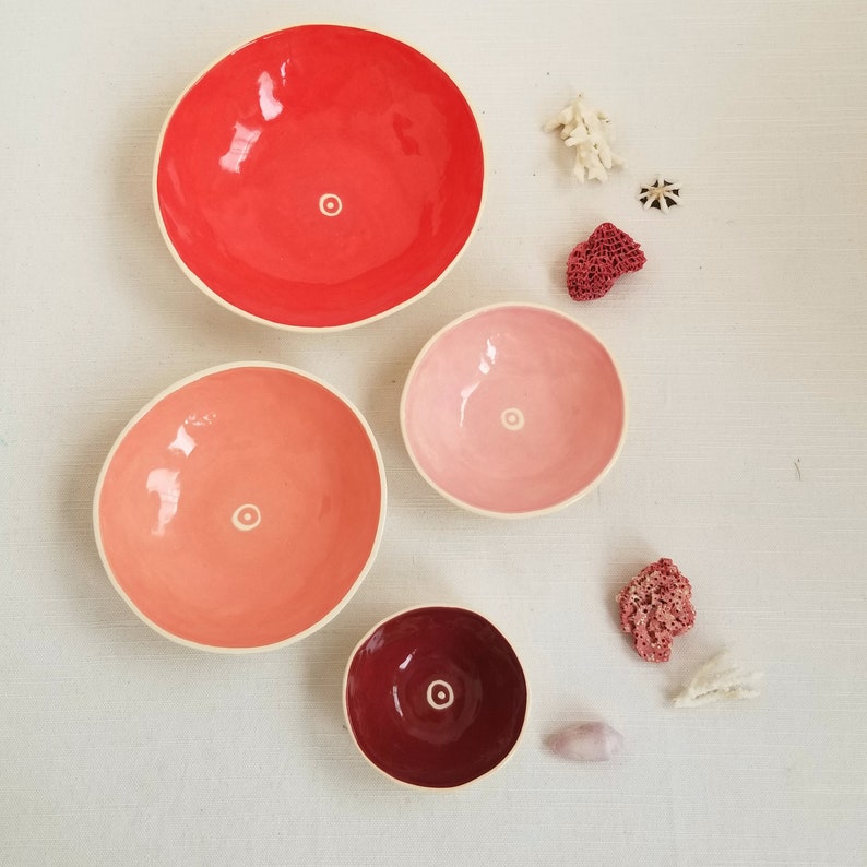 small, handmade ceramic bowls in red, peach, pink& maroon.
white circle and dot in bottom of each, Shown with red & white corals