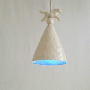 Può includere: Lampada a sospensione in ceramica bianca con un paralume a forma di cono e una piccola statuetta di cavallo in ceramica bianca in cima. L'interno del paralume è di colore blu chiaro.
