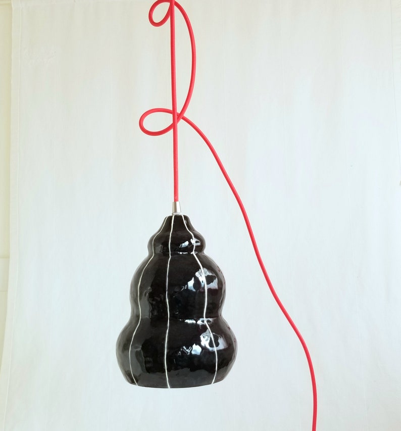 Black ceramic pendant light has an organic form and is detailed with thin white stripes. Hangs from a red 15' plugin cord