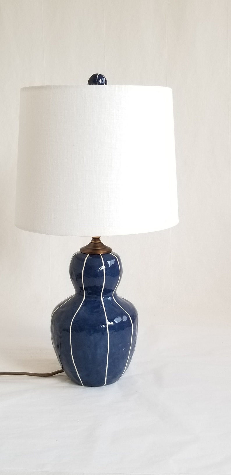 Bedside table lamp in dark blue with thin white stripes. Topped with a white linen shade and matching finial