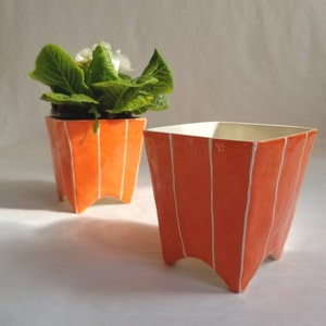 2 Small square ceramic planters in orange with 4 pointed feet. Shown with potted plant and without