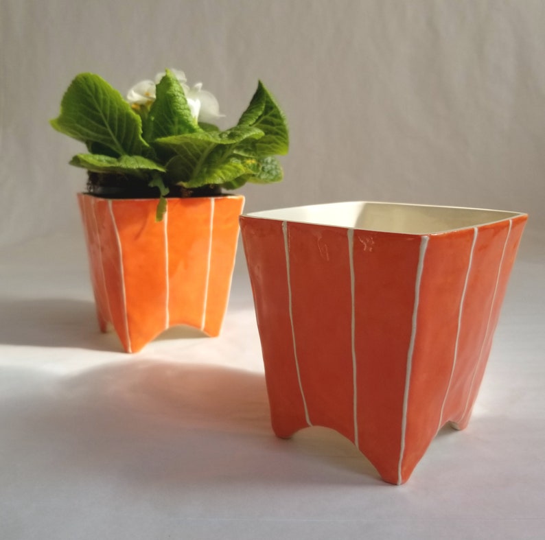 2 Small square ceramic planters in orange with 4 pointed feet. Shown with potted plant and without