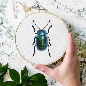 May include: A cross-stitch embroidery hoop featuring a pixelated beetle design in shades of teal, green, and black. The embroidery is set against a white fabric, held within a wooden hoop. The background has a floral pattern.