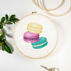 May include: A cross-stitch embroidery project featuring a stack of three colorful macarons in yellow, pink, and teal, centered within a round wooden embroidery hoop. The design is stitched onto white fabric, with a metal tension screw at the top.