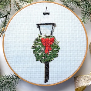 May include: Cross-stitch hoop art depicting a winter scene with a snow-covered lamp post decorated with a green wreath and a red bow. The artwork is set against a light blue background, framed in a wooden hoop, and has a festive holiday theme.