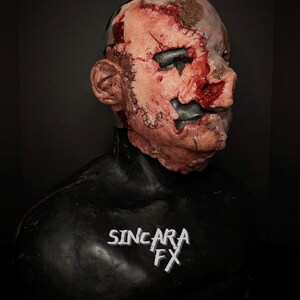 May include: A horrifying mask with a flesh-toned face, showing stitched-up wounds and dark eye sockets. The mask has a black neck piece and the text "SINCARA FX" in white.