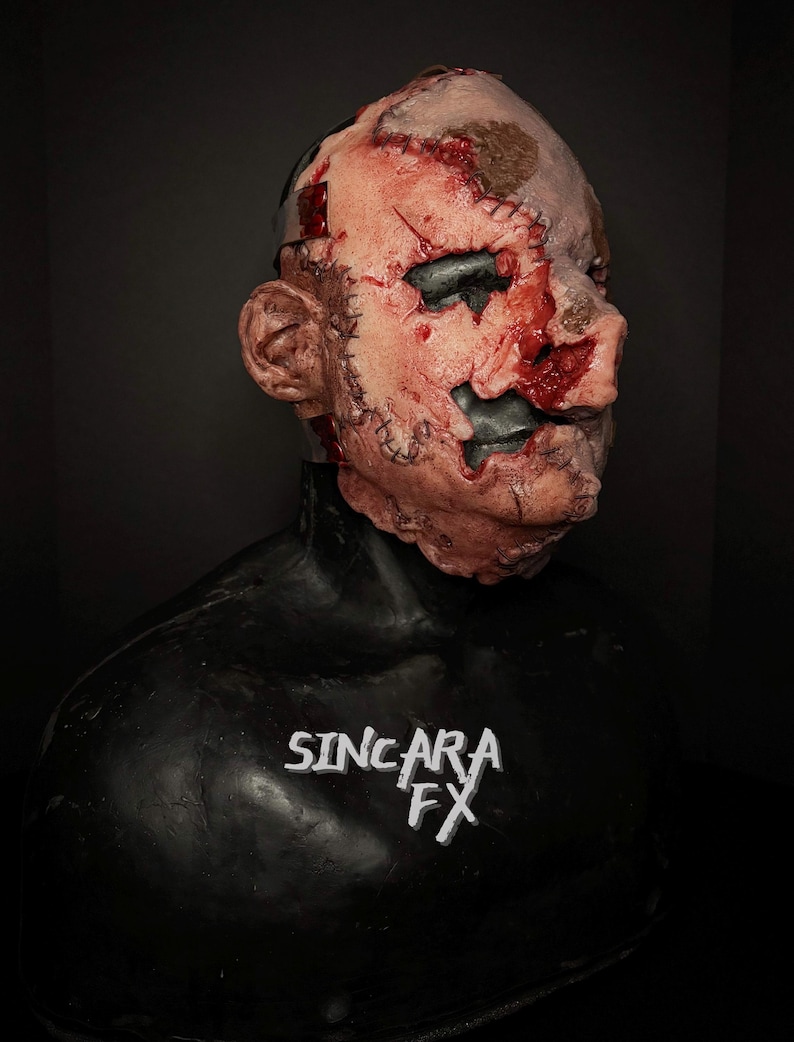 May include: A horrifying mask with a flesh-toned face, showing stitched-up wounds and dark eye sockets. The mask has a black neck piece and the text "SINCARA FX" in white.